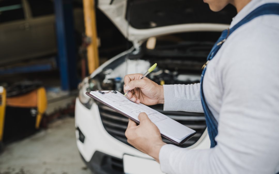 Mechanic holding a clipboard and writing notes in front of a car with its hood open in a garage.